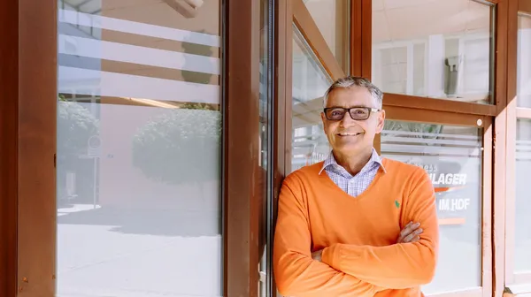 Person mit Brille und orangefarbenem Pullover steht lächelnd vor einer Glasfront in der Sonne.