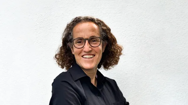 Porträt einer lachenden Person mit Brille und dunklem Hemd vor einer weißen Wand.