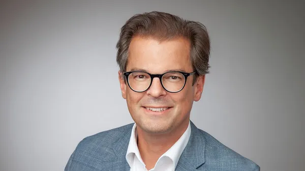 Porträt eines Mannes mit Brille, blauem Sakko und weißem Hemd vor grauem Hintergrund, freundlich lächelnd.