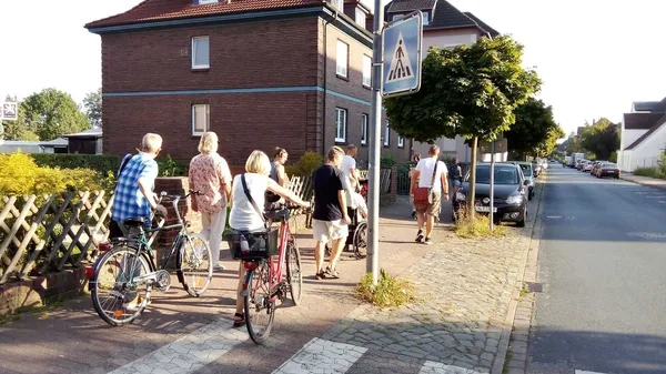 Gruppe von Menschen mit Fahrrädern und Rollstuhl auf einem Gehweg entlang einer Wohnstraße bei sonnigem Wetter.